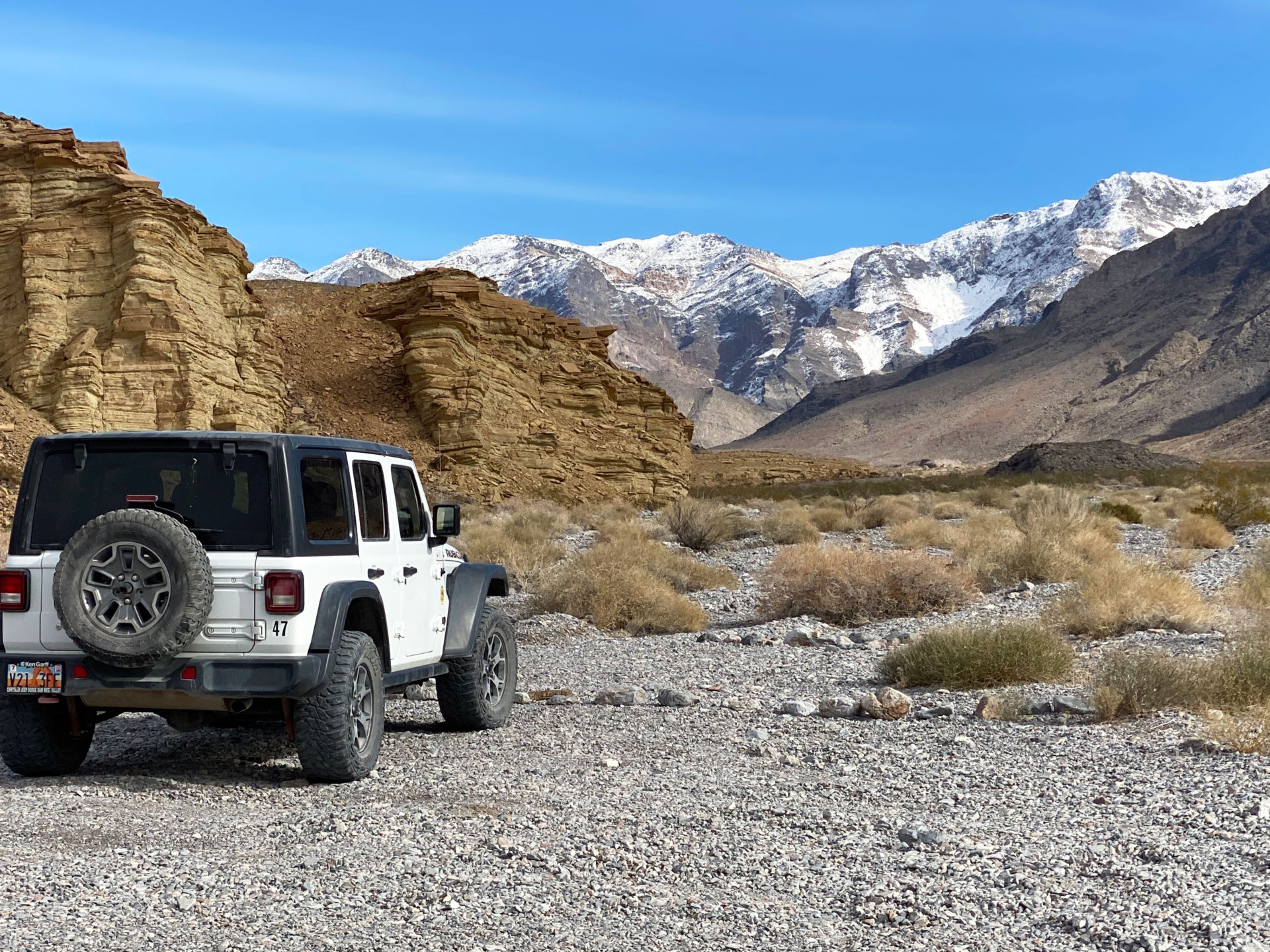 A white Jeep Rubicon parked in front of the camera while Death Valley's surreal and stark desert landscape characterized by vast salt flats, towering sand dunes, and rugged mountains is seen in the background