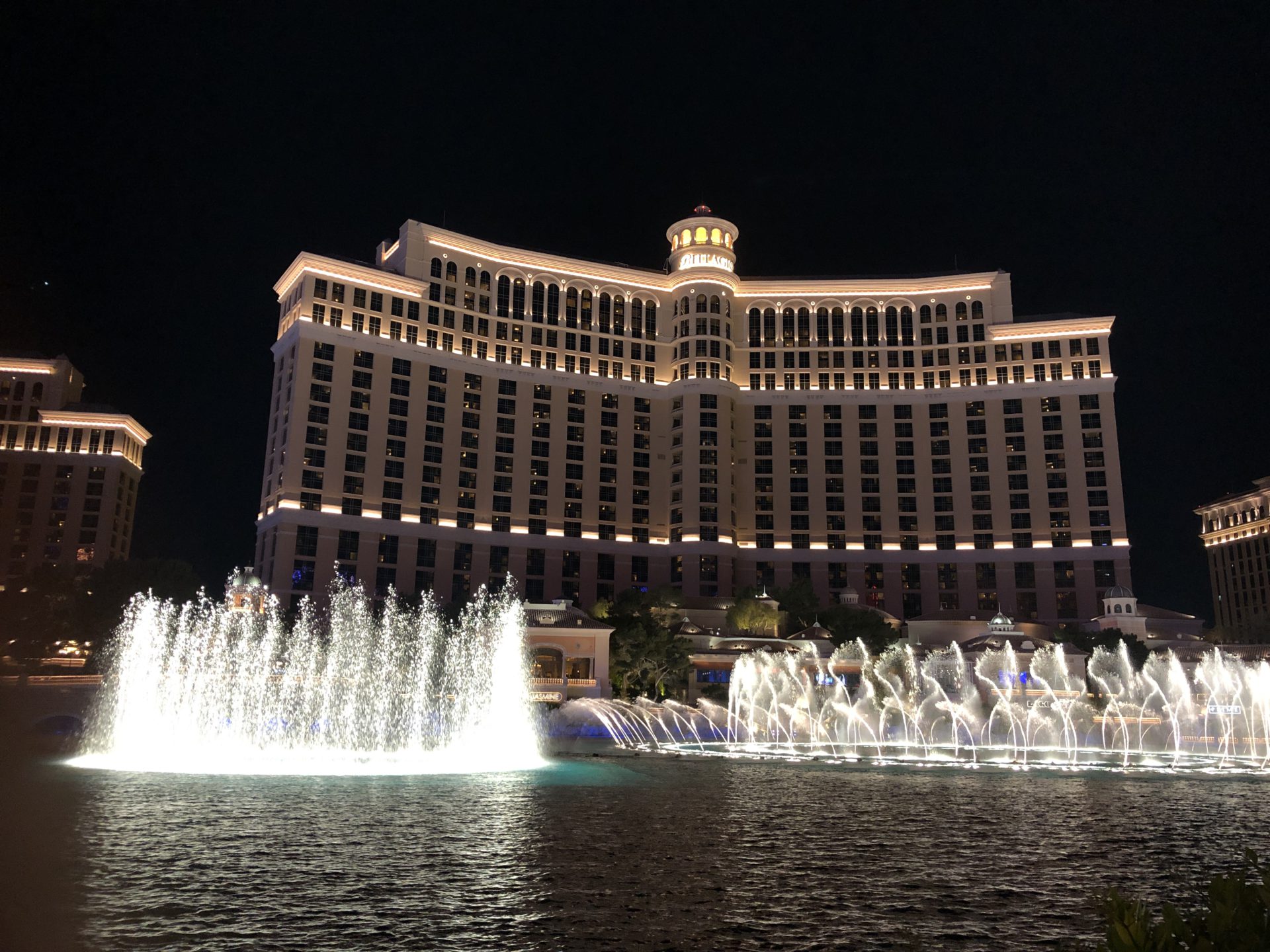The Fountains of Bellagio at night present a display of illuminated water jets against the backdrop of a black Las Vegas skyline