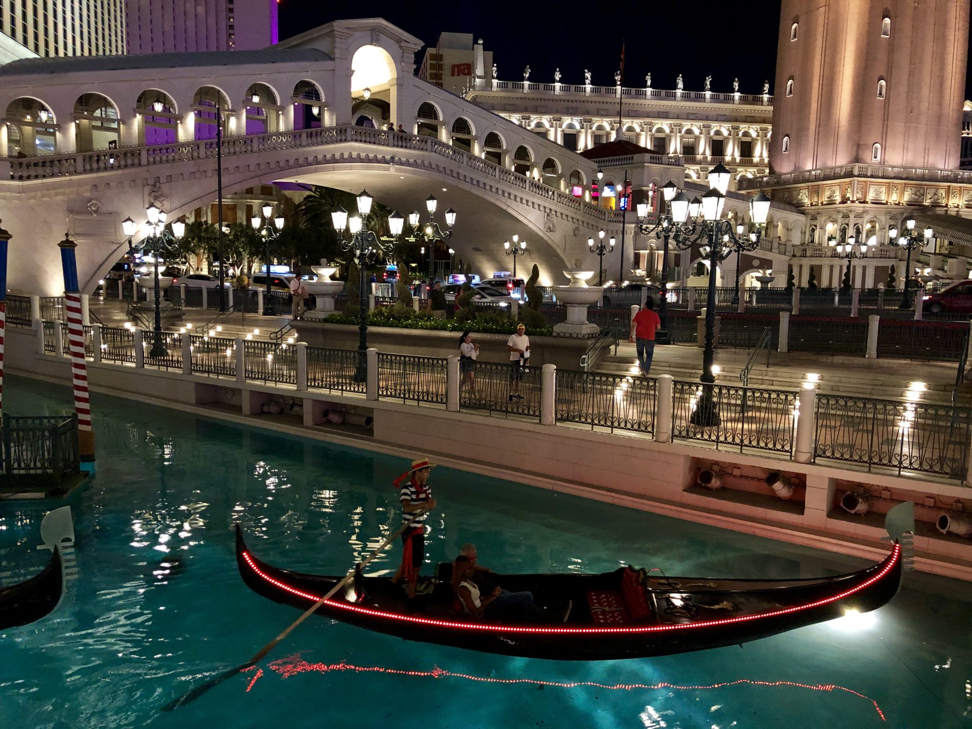 The Venetian Gondola Ride at night with softly lit canals reflecting the shimmering lights of surrounding architecture, creating a magical ambiance reminiscent of Venice under the stars.
