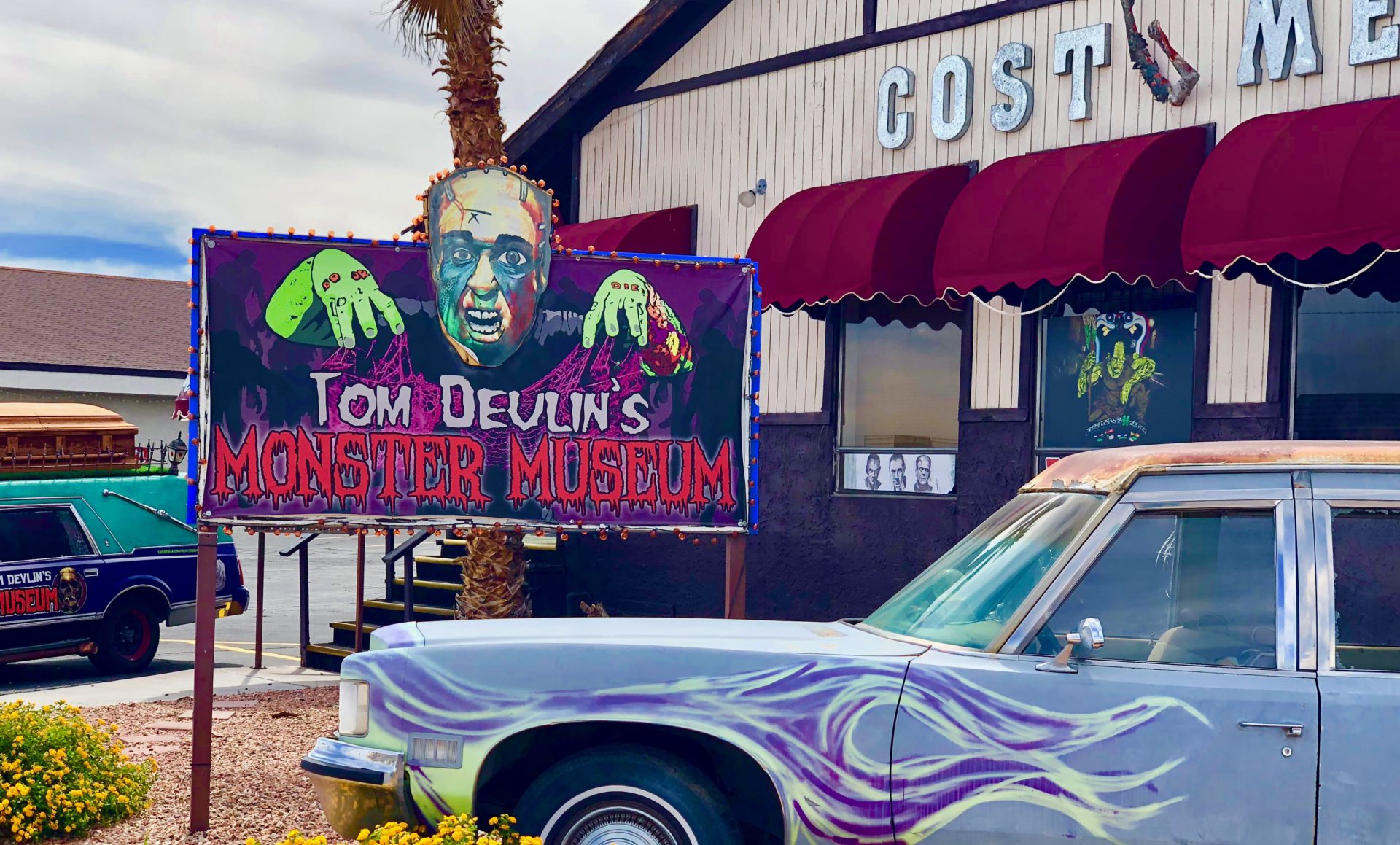 The outside of the Tom Devlin’s Monster Museum where it has a zombie sign saying it's name and a blue retro car next to it. The museum showcases a diverse array of meticulously crafted monster sculptures and memorabilia within a themed environment reminiscent of classic horror films.