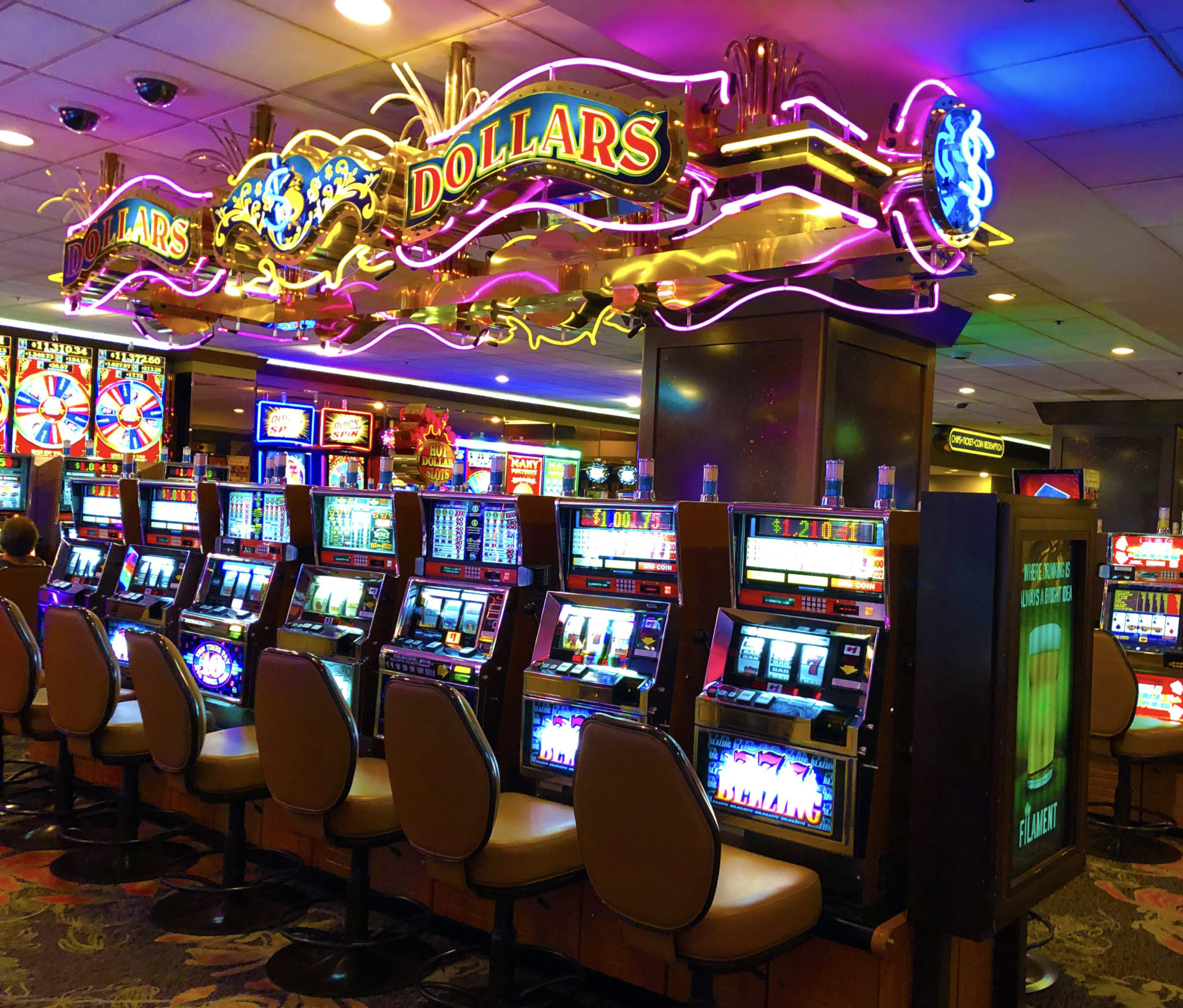 A view of a Las vegas casino where yellow tan seats are lineup to slot machines and a colorful decorative sign hands above saying Dollars Dollars