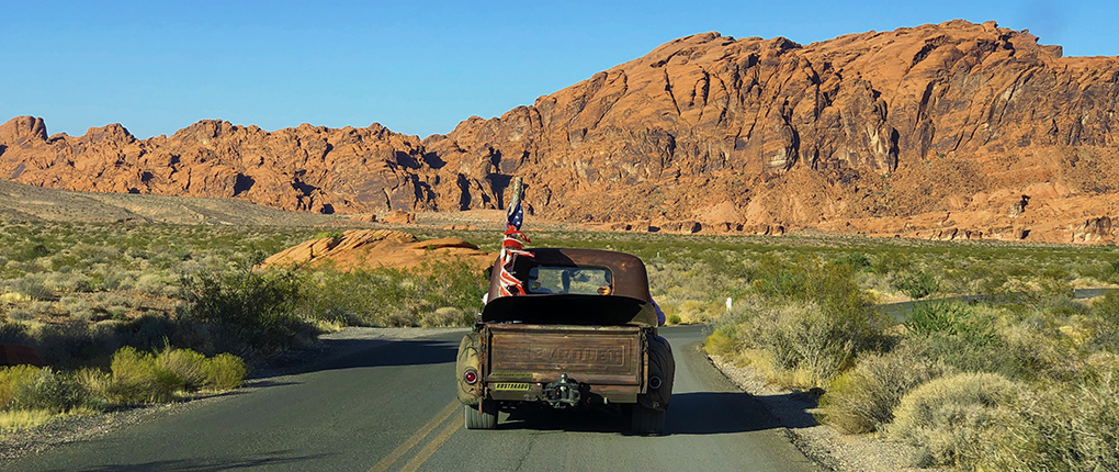 A rustic chevrolet truck driving in the Las Vegas road where clear blue sky, orange hue rocky mountains, and greenery are seen in the background.